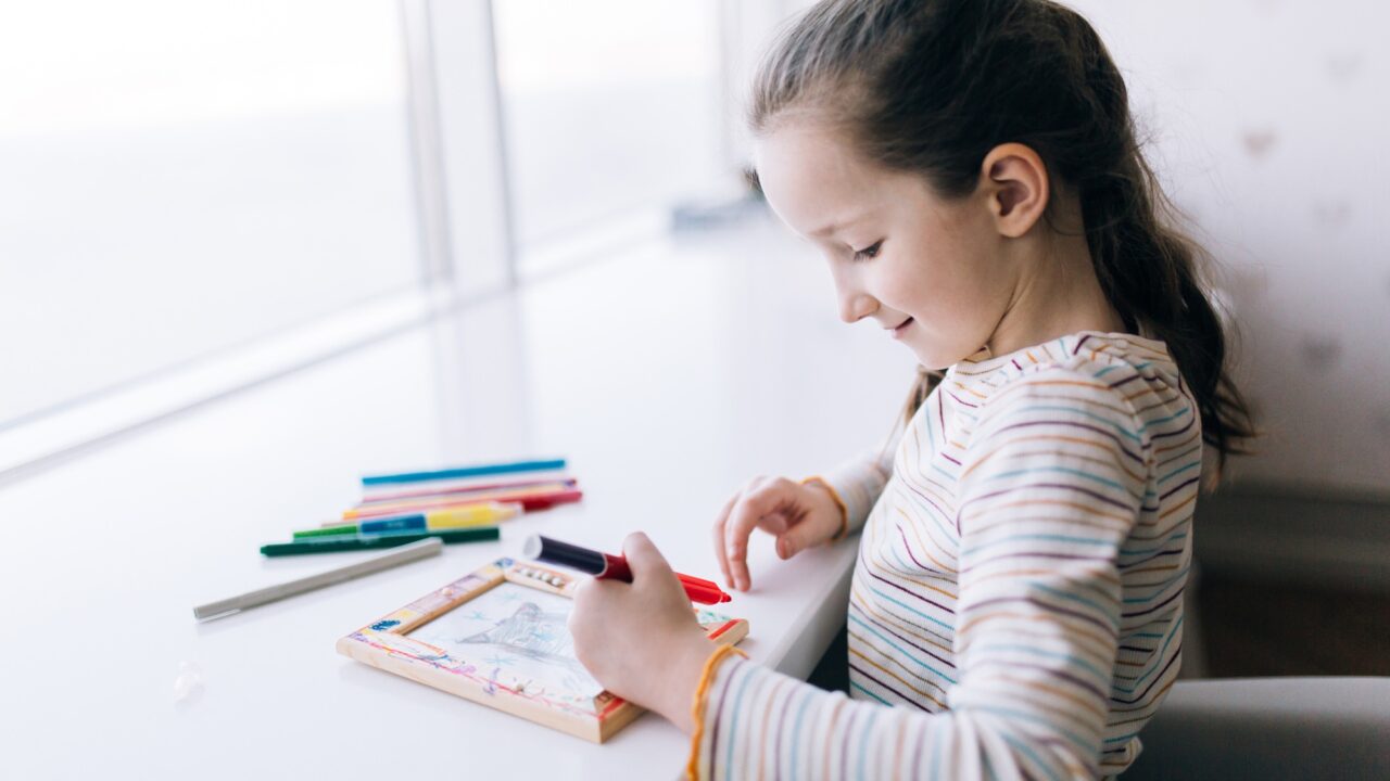 A girl sits at a table and draws, decorates a frame for a photo with drawing, children creativity