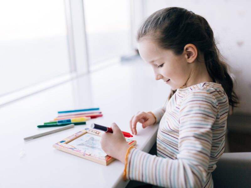 A girl sits at a table and draws, decorates a frame for a photo with drawing, children creativity