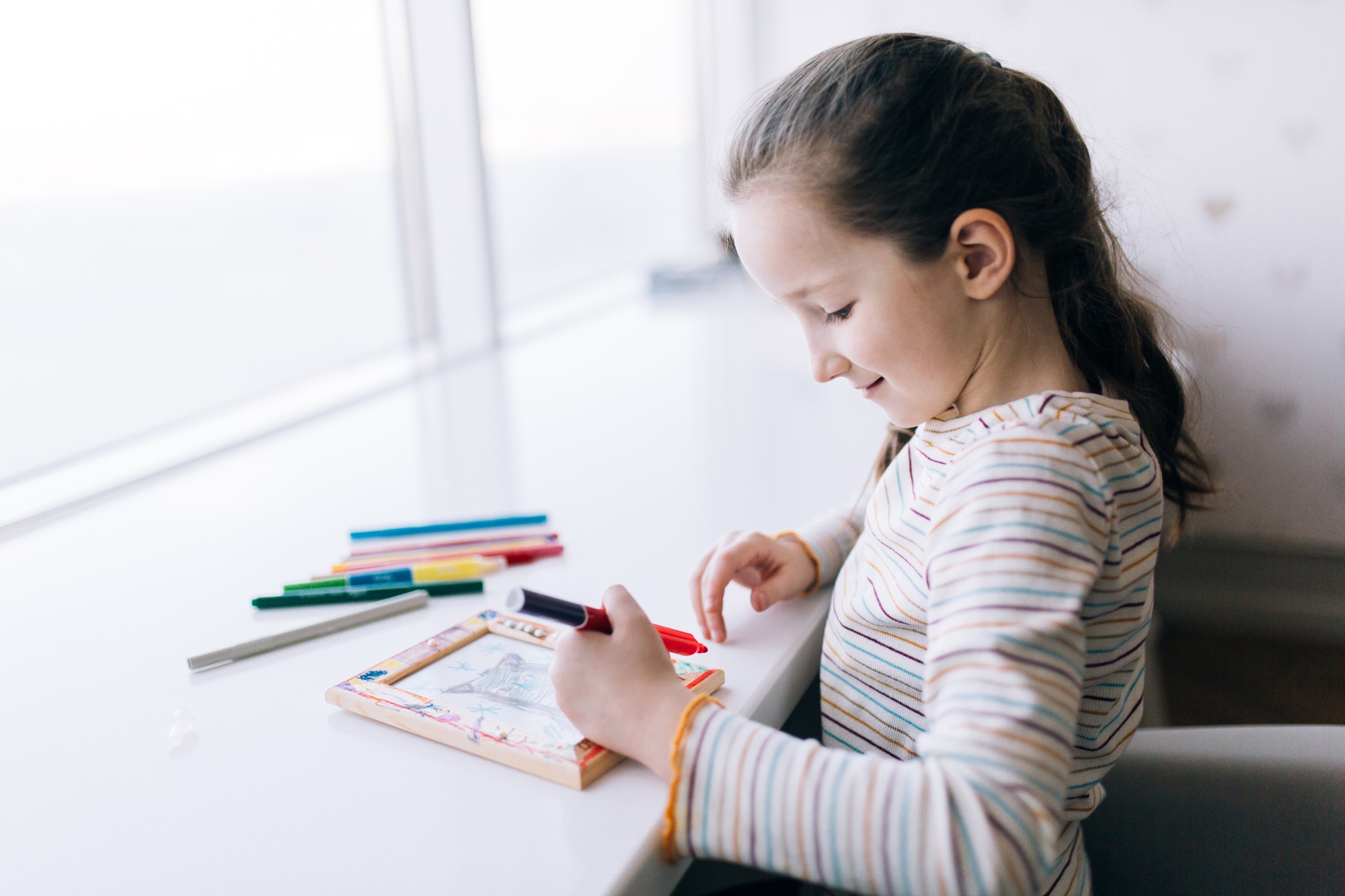 A girl sits at a table and draws, decorates a frame for a photo with drawing, children creativity