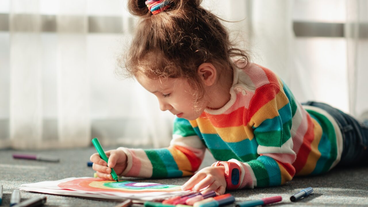 A little girl sits on the floor in the sun and draws a rainbow with markers.