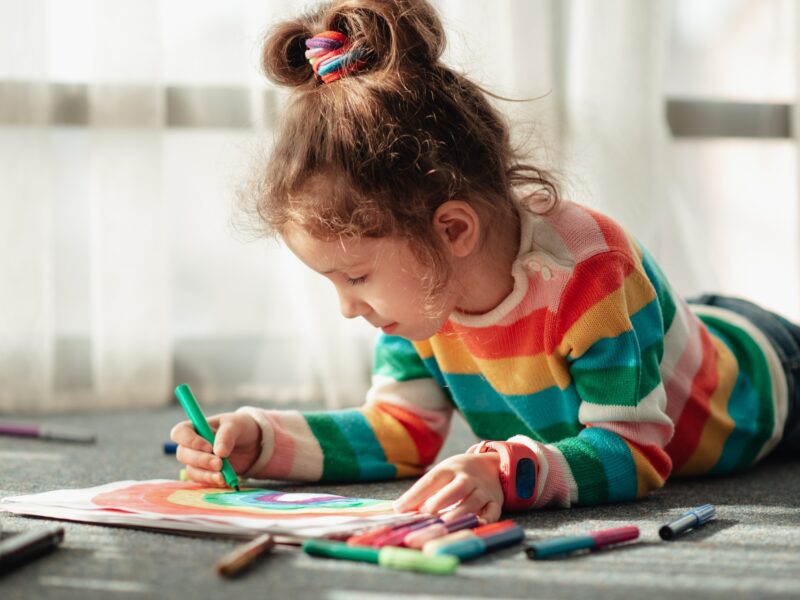 A little girl sits on the floor in the sun and draws a rainbow with markers.
