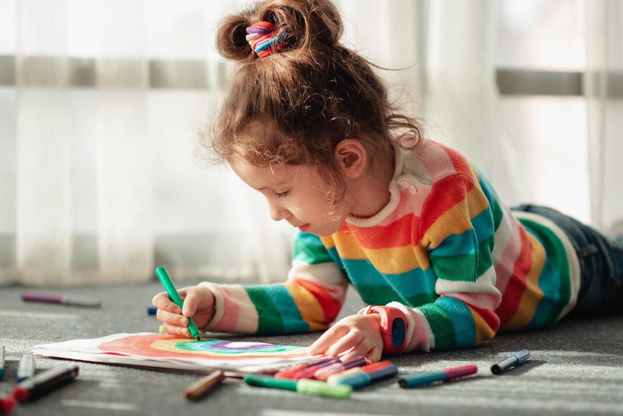 A little girl sits on the floor in the sun and draws a rainbow with markers.