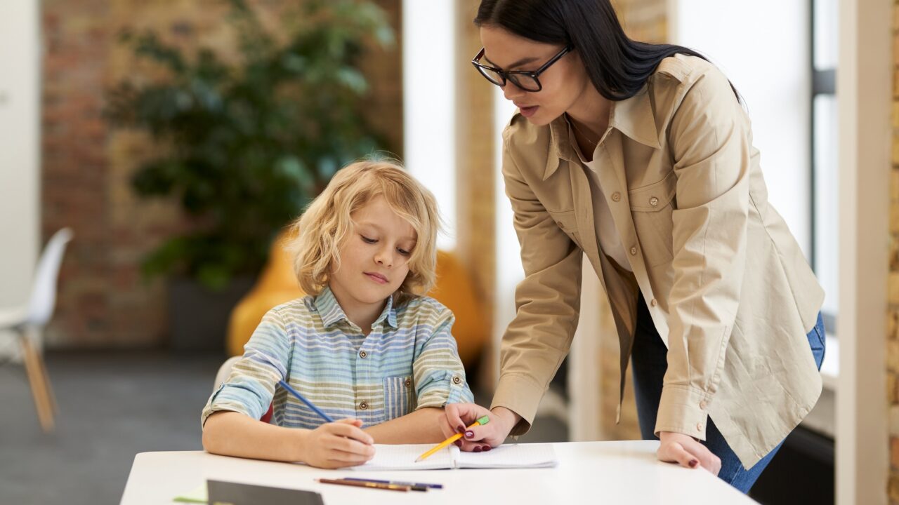 Supportive young female teacher in glasses helping little boy with the task. Kid studying in