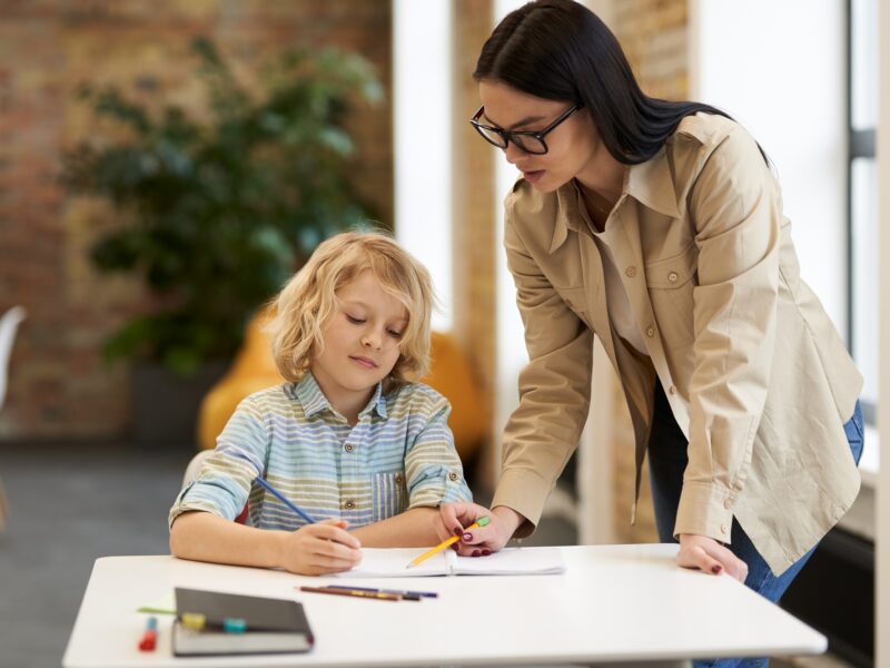 Supportive young female teacher in glasses helping little boy with the task. Kid studying in