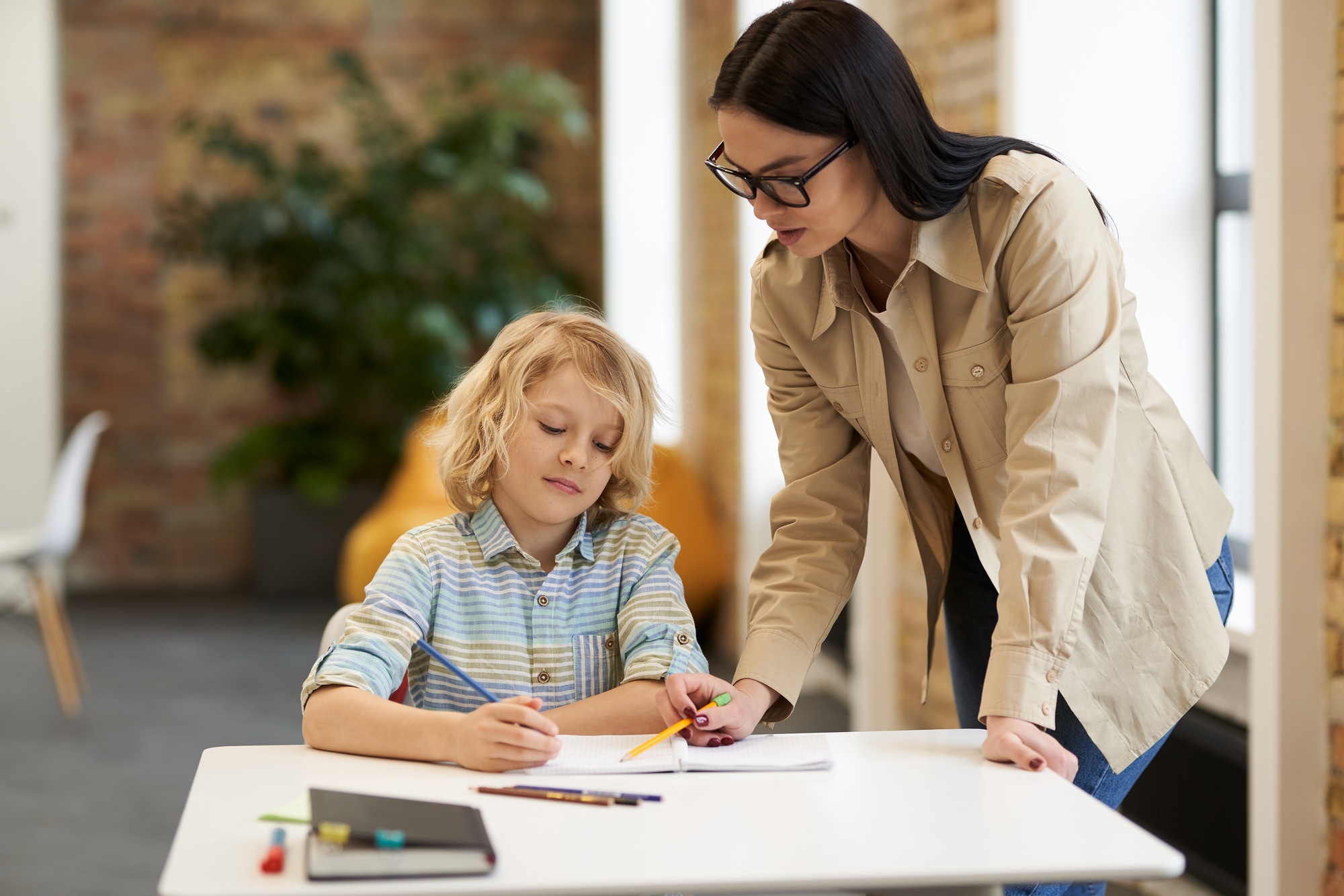 Supportive young female teacher in glasses helping little boy with the task. Kid studying in