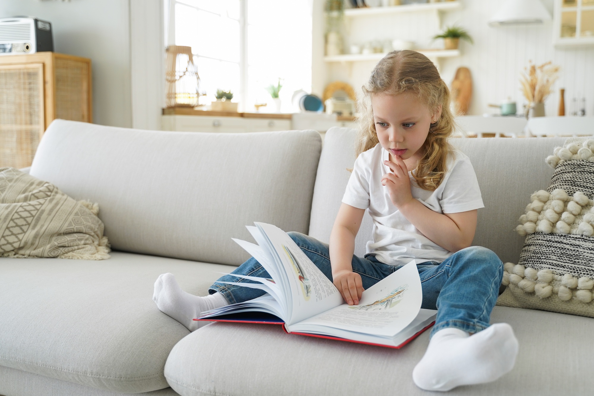 Pensive little child girl preschooler turns pages of fairytale book for reading, sit on sofa at home