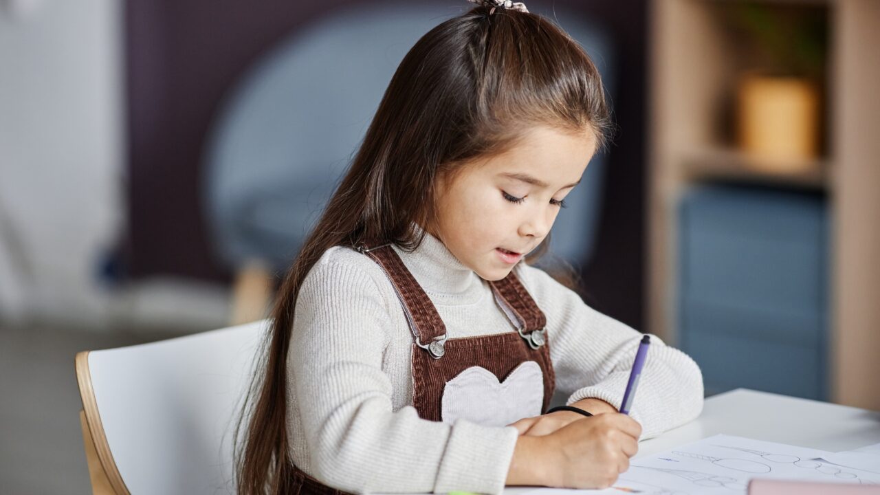 Diligent little girl with dark long hair drawing with crayons