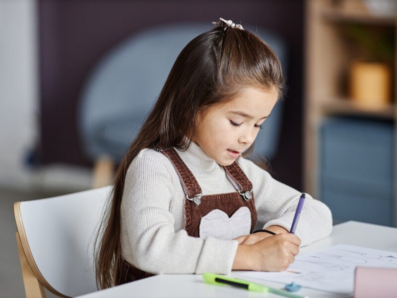 Diligent little girl with dark long hair drawing with crayons